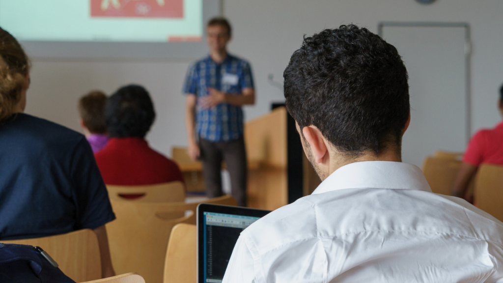 A student with dark hair and a white collared shirt sitting in front of a laptop in a seminar room. A lecturer stands in the blurry background and hosts a lecture with a PowerPoint.