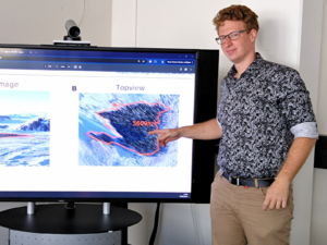 The PHD Student and biophysicist Alexander Winterl standing next to a screen, pointing at a satellite picture with a colony of penguins.