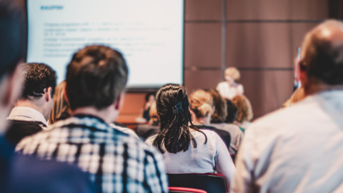 A group of students sitting in a classroom. In the foreground you can see the backs of the audience's heads, in the blurred background you can see a lecturer giving a presentation.