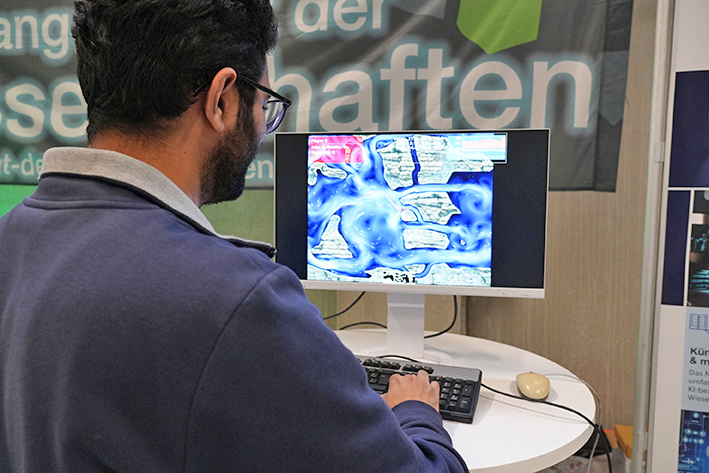 The photo shows a person facing a monitor displaying a colorful screen that appears to show a game or simulation involving a blue, fluid-like environment with floating rock structures. The person is typing or controlling the display using a keyboard and mouse. The setting appears to be an indoor science fair.