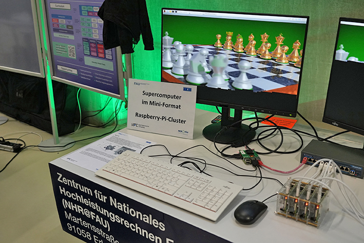 The photo shows a computer demonstration setup on a table. A monitor displays a 3D chessboard with gold and silver chess pieces. In front of the screen, there is a white keyboard, a black mouse, and several cables connected to a small cluster of Raspberry Pi boards stacked together on the right side of the table. The overall setup appears to demonstrate a miniature supercomputer or parallel computing cluster built with Raspberry Pi devices.