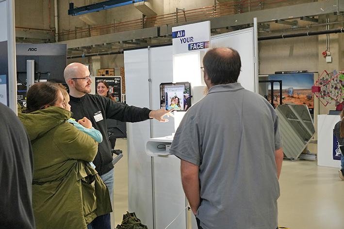 The photo shows a small group of people gathered around a booth at an indoor event. One person, standing near a display labeled “AI YOUR FACE”, appears to be explaining or demonstrating something on a tablet or screen mounted on a stand. The screen shows a person’s face, likely part of a facial recognition or image-processing demo. The setup suggests a technology or science fair focused on artificial intelligence or digital innovations.