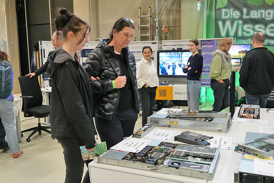 This image captures a group of adults gathered around a display of technical hardware, likely at a university event, given the visible signage and logo. The setting suggests a focus on technology, education, or research, with participants engaged in discussing or learning about the presented hardware.