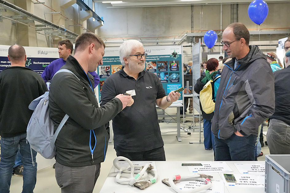 This image captures a focused discussion between three men at an indoor university exhibition, likely involving hardware or specialized equipment. There are several informational posters and banners visible, one of which features the FAU (Friedrich-Alexander-Universität) logo and discusses computer projects. The overall impression is one of serious technical engagement at a public-facing event.