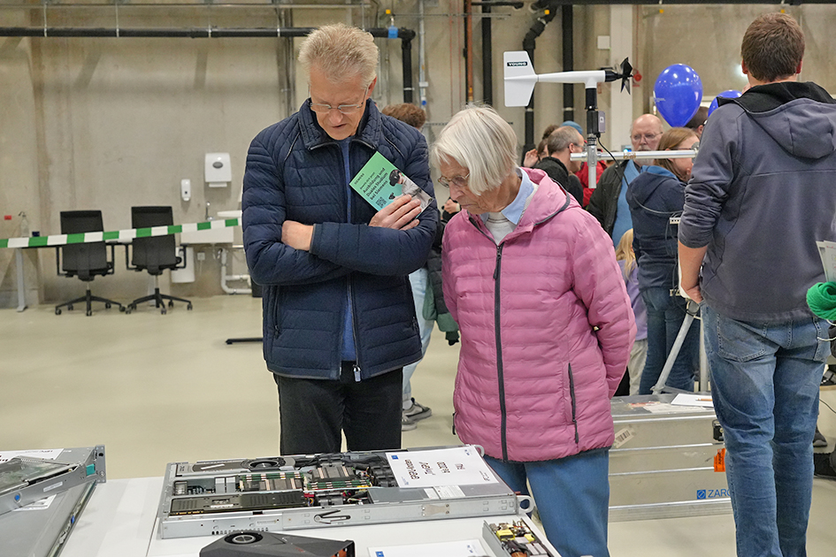 The image shows the interaction between an older man and an older woman examining technical equipment on a table, presumably as part of an open day at a university. The scene conveys a sense of public interest and engagement with technology.