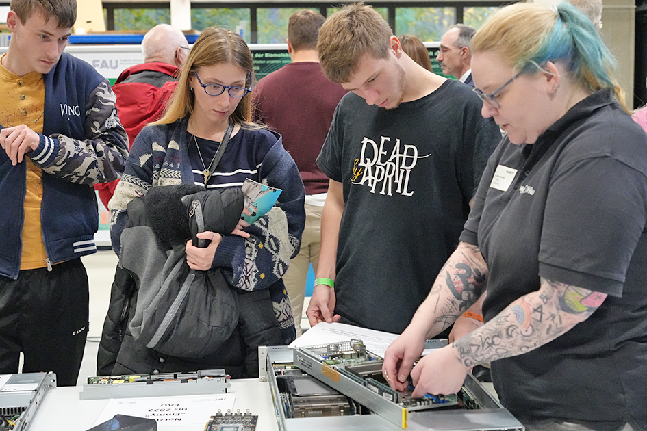 This image captures a close-up view of a technical demonstration or presentation, likely at a university open house, with an instructor and several younger attendees focused on computer hardware. The overall atmosphere is one of focused instruction and learning about technical hardware.