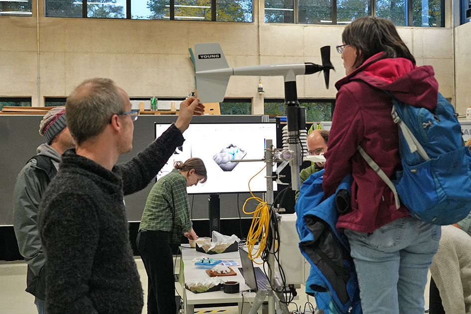 This image captures a group of adults gathered around a demonstration that combines physical models, digital displays, and scientific equipment, likely at an educational event. The overall scene conveys an interactive, hands-on learning experience in a technical or scientific field, involving both physical and digital models.