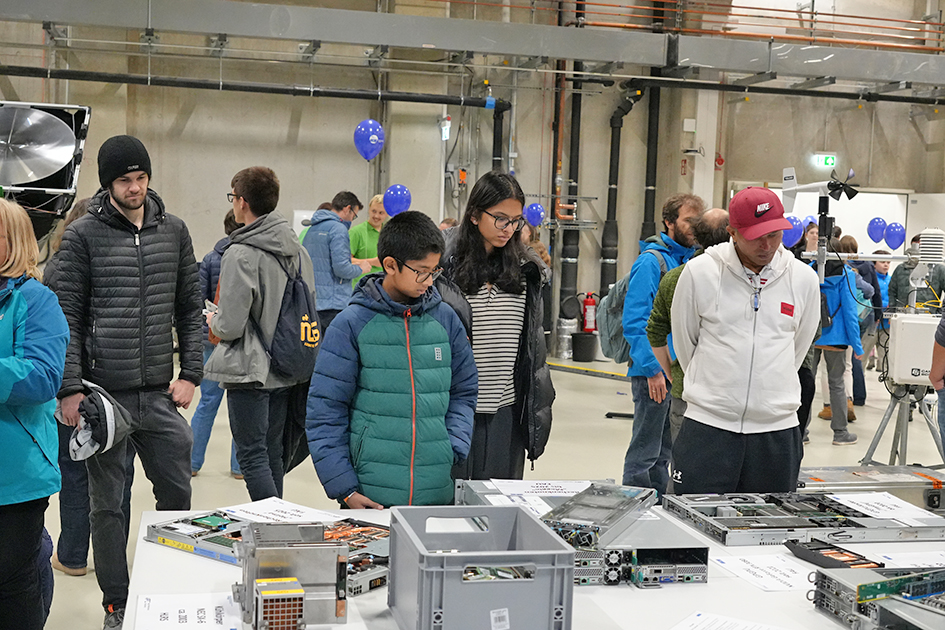This image captures a group of people, including adults and minors, gathered around a display of computer or server hardware in a large, industrial indoor space, likely at an educational event. The image conveys a sense of public engagement with technology and research in a functional, laboratory-like environment.