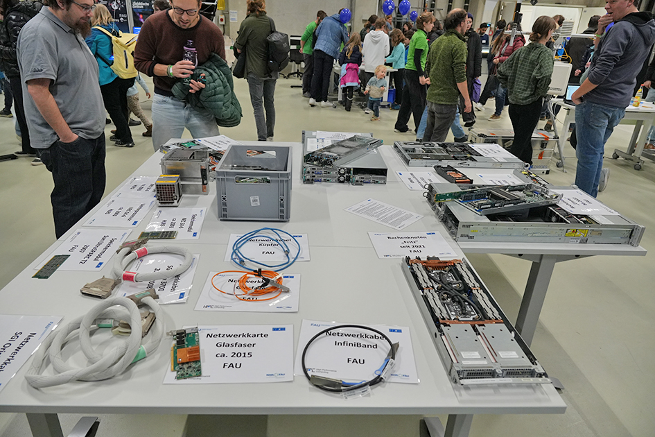 This image focuses on a display table featuring various computer and networking hardware components, surrounded by a crowd of people in a large indoor exhibition space. The central element is a large white table covered with technical equipment. The display includes several open server chassis, showcasing internal components like circuit boards and drives. There are also smaller hardware modules, a grey plastic storage box, and various networking cables and components. Numerous people are visible surrounding the table and filling the background, suggesting a busy public event. The overall impression is a detailed, hands-on demonstration of specialized computer networking and server technology.