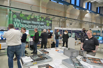This image captures a wide-angle view of a busy scientific exhibition taking place in a large, industrial hall. The focus is on a hardware display table and several groups of people interacting. A long white table dominates the lower half of the image, showcasing various computer components, primarily open server chassis. Small signs and cables are scattered among the hardware. Numerous people are visible, indicating a high level of attendance. The overall atmosphere is one of lively public engagement at a well-attended science and technology event.