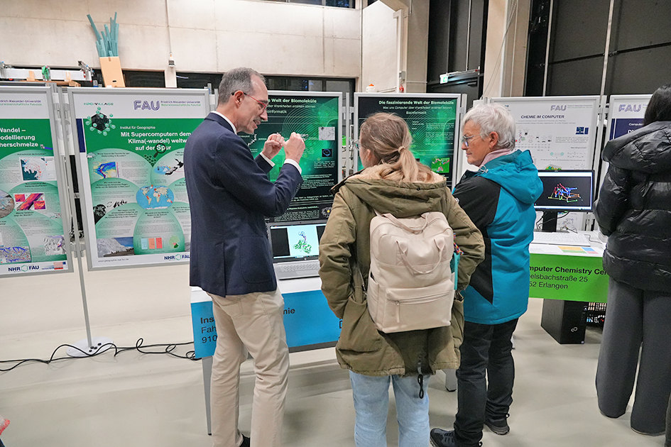 This image captures a focused discussion taking place in front of a series of scientific and technical posters, suggesting an academic exhibition setting. A man is presenting information to two women. The group is gathered around a table with a laptop, a monitor, and several large posters. The scene conveys a sense of informed discussion and intellectual engagement within a university or research context.