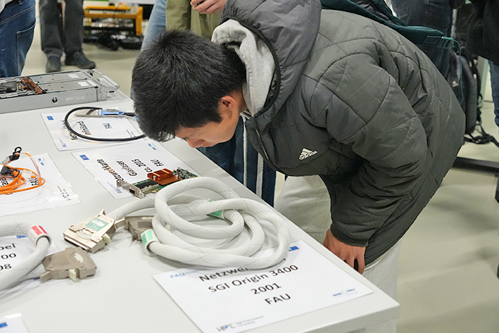 This photo shows a young person intently examining cables and hardware on a table. In the foreground lies a thick bundle of white, coiled cables with connectors, next to a sign that reads: 