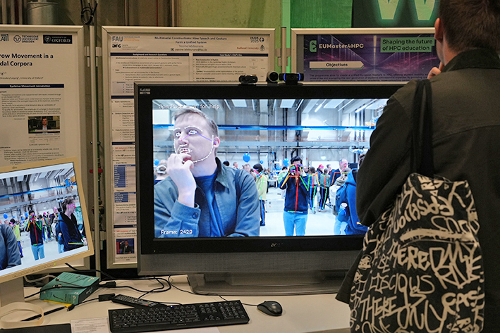 This image captures a close-up view of a technology demonstration focused on human movement and tracking, likely at a research exhibition. The main screen shows a live or recorded video feed of a man's face overlaid with white tracking points and lines, forming a mesh or skeleton around his eyes, mouth, and jawline. This indicates real-time facial motion capture or analysis. The scene clearly showcases a project involving computer vision, data analysis, and human-computer interaction, specifically the tracking and analysis of facial and body movement.