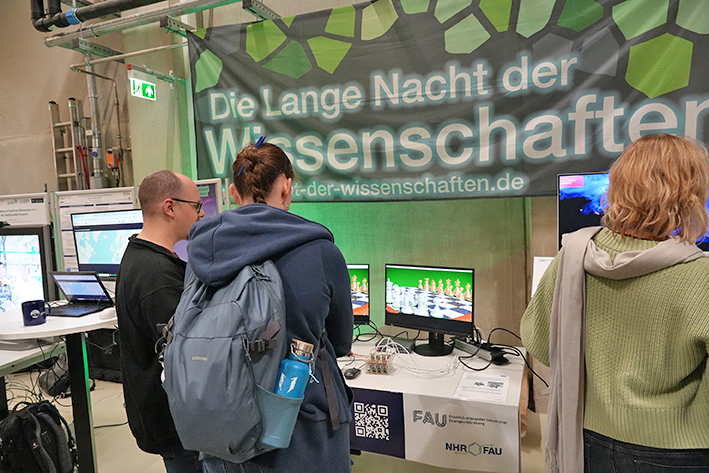 This image captures a view of attendees at a public science event, focusing on a display table featuring computer monitors and a large overhead banner. Three adults are gathered around a table with computer equipment. The overall atmosphere is one of public curiosity and engagement with scientific and technological research.