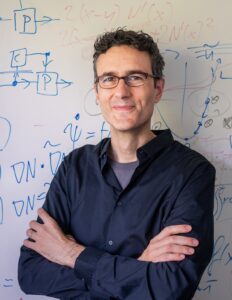 A man with short curly hair and glasses stands smiling with his arms crossed in front of a whiteboard covered in handwritten mathematical equations and diagrams.