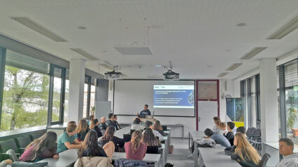 The image shows a lecture setting. A presenter is standing at the front of the room, speaking to a group of students. Behind the presenter, there is a large screen displaying a slide, likely part of a presentation. The students are seated at long tables arranged in rows, facing the front.